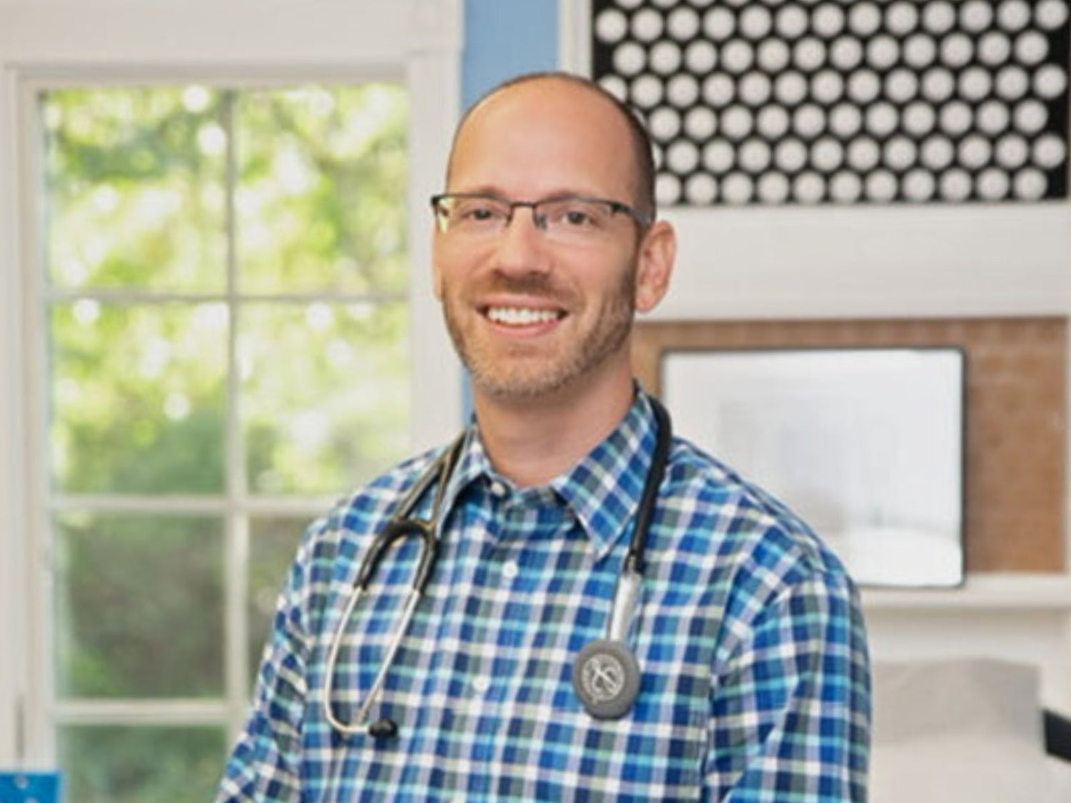 Doctor wearing a blue checkered shirt with a stethoscope, standing in a room with a window and decor.