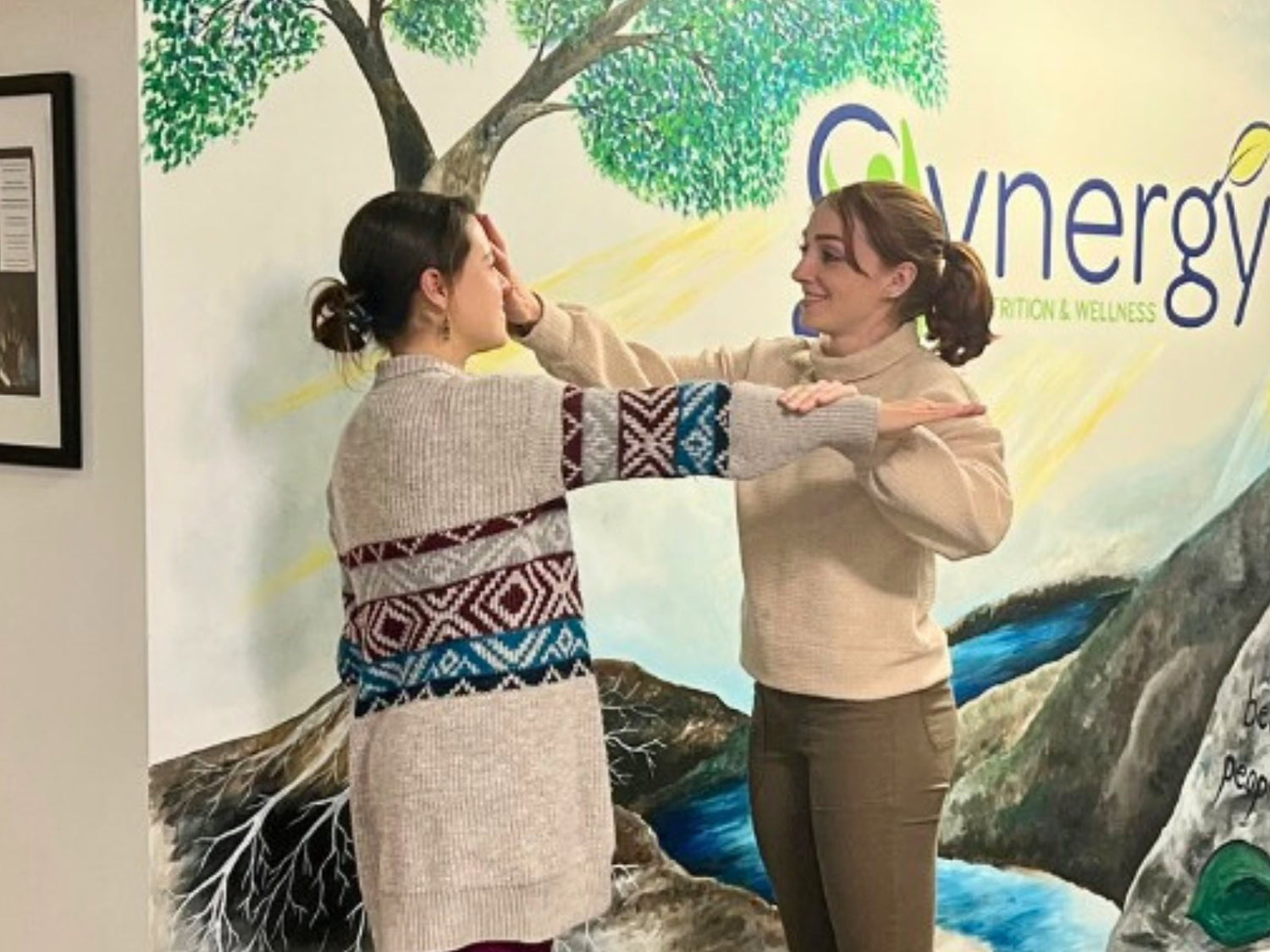 Two women interacting in front of a mural with Synergy branding