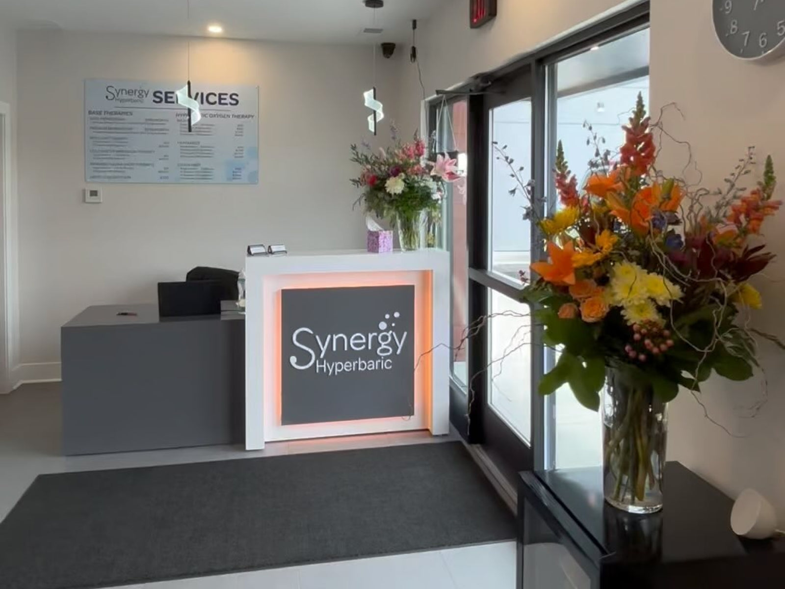 Reception area with 'Synergy Hyperbaric' sign, flowers, and a clock on the wall  at Synergy Nutrition and Wellness in Western New York.