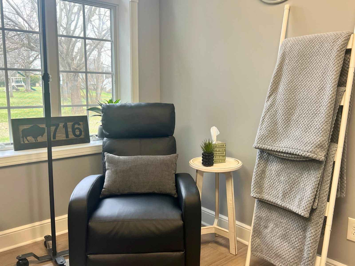 Black recliner chair with gray pillow and blanket in a room with a window and small table.